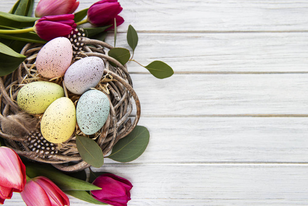 Easter background with colorful eggs in nest and pink tulips on a white wooden background. Top view with copy space - 写真・画像