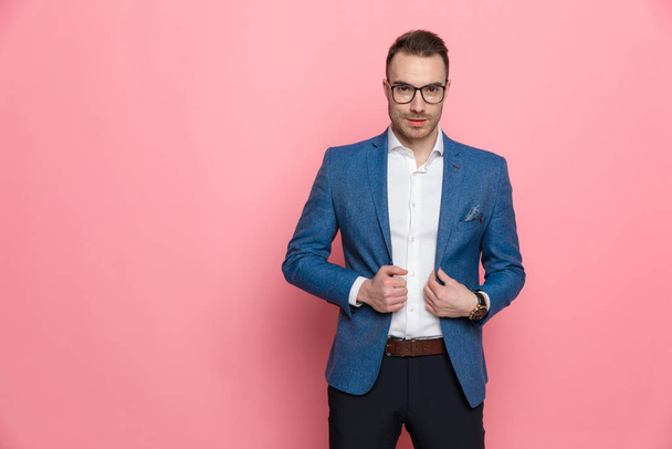 Gemotiveerde slimme casual man aanpassen van zijn jas en glimlachen tijdens het dragen van een bril en staan op roze studio achtergrond - Foto, afbeelding
