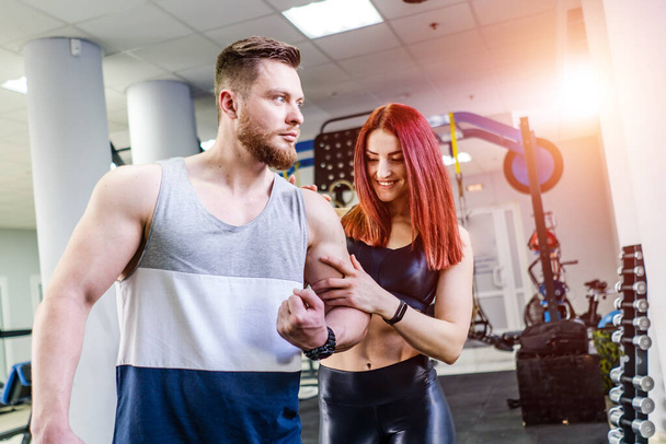 Strong athlete showing muscles to beautiful woman in fitness club. Young woman is touching male's biceps on the bend arm. - Фото, зображення