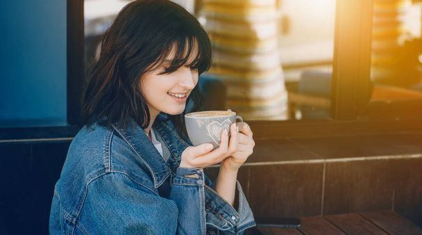 Smiling caucasian girl with short hair and blue jacket is drinking a coffee outside near a coffee shop - Photo, Image