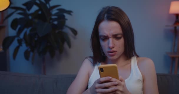 Close up of young woman sitting on sofa with smartphone looking at screen and feel sad. Pretty girl covering face with hand and crying after reading message about relations break up . - Πλάνα, βίντεο