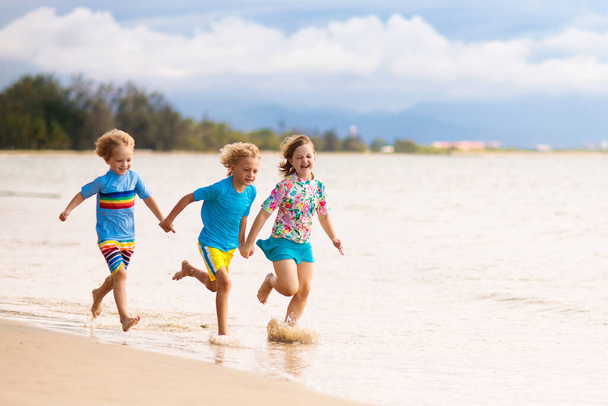 Παιδιά που παίζουν σε τροπική παραλία. Τα παιδιά κολυμπούν και παίζουν στη θάλασσα στις καλοκαιρινές οικογενειακές διακοπές. Η άμμος και το νερό διασκέδαση, προστασία από τον ήλιο για το μικρό παιδί. Αγοράκι και κορίτσι τρέχουν και πηδάνε στη θάλασσα.. - Φωτογραφία, εικόνα