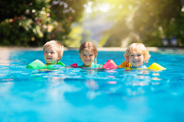 Gyerekek játszanak szabadtéri medence trópusi üdülőhely. Úszó segédeszköz kisgyereknek. A baba búvárkodni tanul. Egy csapat gyerek játszik a vízben. Színes mentőmellény. Strand és nyári szórakozás. - Fotó, kép