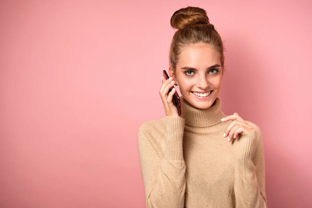 A girl in a turtleneck with a high beam on her head brings the phone to her ear and smiles on a pink background. - Photo, Image