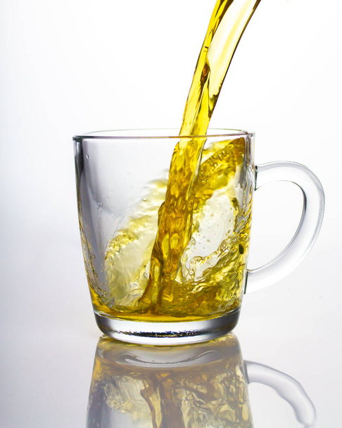 Strumień żółtej zielonej herbaty płynącej do przezroczystego szklanego kubka, piękny spray latający w łuku, odbicie. Herbata nalewana - Zdjęcie, obraz
