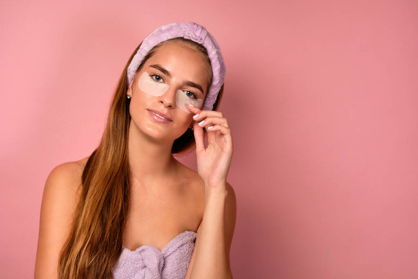 A girl with clean skin with a bandage on her hair stands on a pink background and corrects patches under her eyes - Foto, Imagen