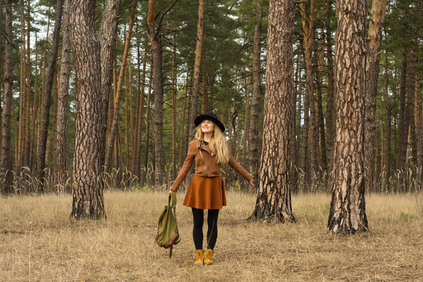 Sonbahar ormanında sırt çantası ve şapkasıyla yürüyen bir kız. Eski tarz. Soğuk giysi takımı. Macera, turizm, yürüyüş, seyahat ve insan konsepti.  - Fotoğraf, Görsel