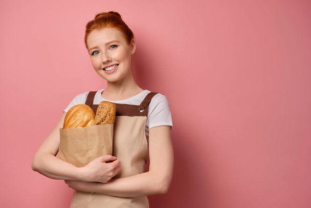 A cute redhead girl with a high bun in a apron stands on a pink background smiling broadly and holding a bag of bread to herself - Фото, изображение