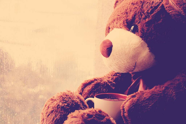 Коричневый игрушечный медведь сидит на окне с чашкой чая и смотрит на капли дождя на стекле
. - Фото, изображение
