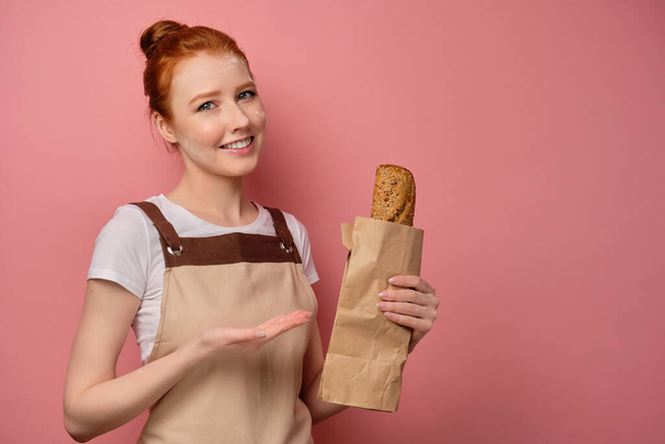 A cute red-haired girl with a high bun in a beige apron, with her face in flour and smiling, points to a packet with a loaf - Фото, изображение