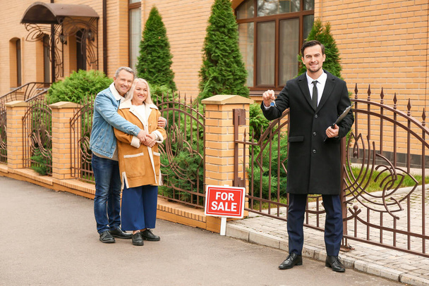 Agente inmobiliario masculino con llave y pareja madura cerca de casa en venta al aire libre
 - Foto, imagen