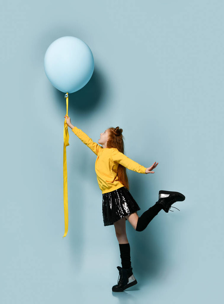Teenage female in yellow sweatshirt, black skirt, knee-highs, boots. She holding balloon, posing sideways on blue background. - Photo, Image