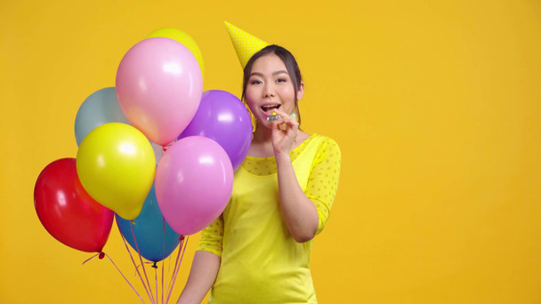 Ασιάτισσα με μπαλόνια φυσάει φυσητήρα κόμμα απομονώνονται σε κίτρινο  - Πλάνα, βίντεο