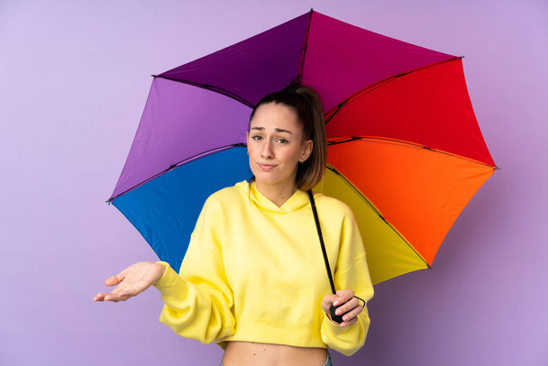 Młoda brunetka trzymająca parasol nad odizolowaną fioletową ścianą robiąc wątpliwy gest podnosząc ramiona - Zdjęcie, obraz
