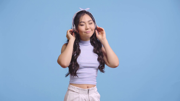 χαρούμενο κορίτσι από την Ασία ακούει μουσική σε ακουστικά απομονωμένα στο μπλε  - Πλάνα, βίντεο