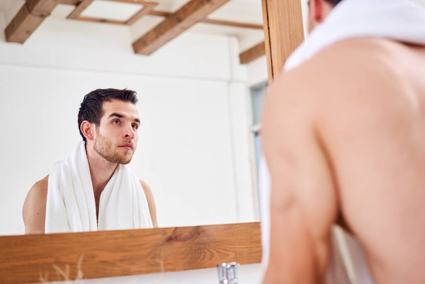 Young male with white towel on shoulder stands near mirror in bath at morning. - Foto, Imagen