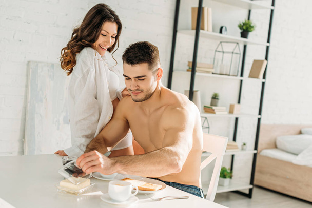 seductive girl in white shirt sitting on kitchen table while sexy boyfriend having breakfast  - Foto, imagen