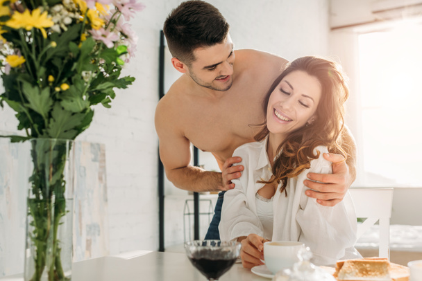 sexy shirtless man hugging happy girl sitting at kitchen table near vase with flowers - Photo, Image