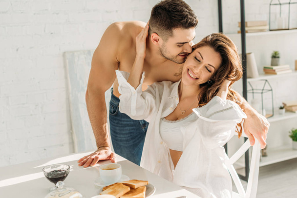 handsome shirtless man kissing smiling girl touching his face while sitting near breakfast - Foto, Imagem