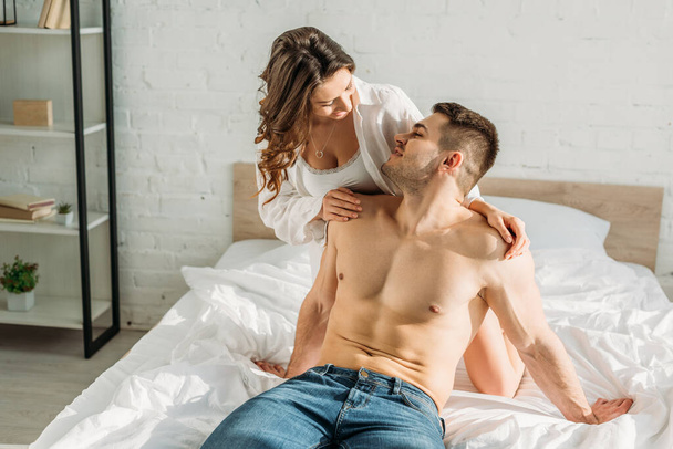 sensueel meisje in wit shirt en bralette kijkend naar shirtloze, sexy man zittend op bed in jeans - Foto, afbeelding