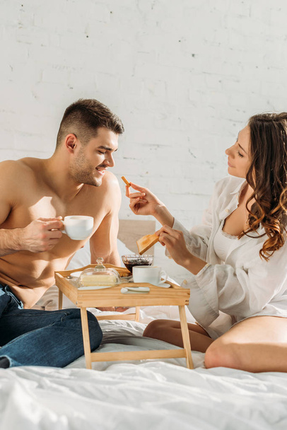 attractive girl giving toast to sexy boyfriend holding cup of coffee near bed tray with tasty breakfast - Foto, Bild