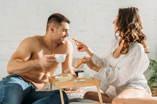 smiling girl giving toast to shirtless boyfriend holding cup of coffee near bed tray with delicious breakfast - Photo, Image