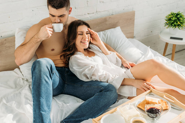 陽気な彼女の近くでコーヒーを飲んでいるハンサムな白人男性とおいしい朝食とベッドトレイ - 写真・画像