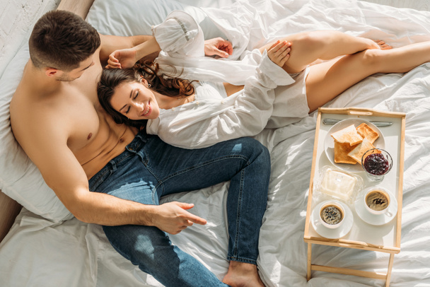 top view of shirtless man in jeans and sexy girl in white shirt lying near bed tray with delicious breakfast - Photo, Image