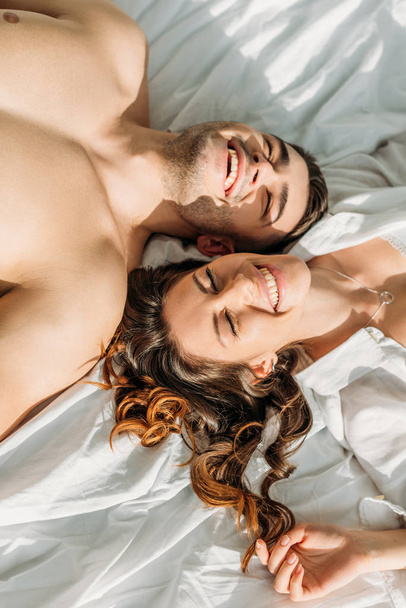 κορυφαία άποψη του χαρούμενου, σέξι ζευγάρι χαμογελά με κλειστά μάτια, ενώ βρίσκονται στο κρεβάτι μαζί - Φωτογραφία, εικόνα