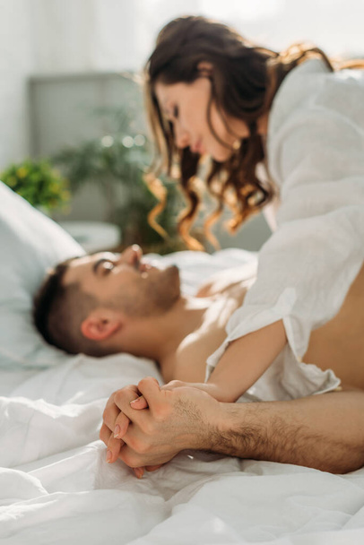 messa a fuoco selettiva di seducente ragazza che si tiene per mano con l'uomo sexy mentre sdraiato su di lui a letto
 - Foto, immagini