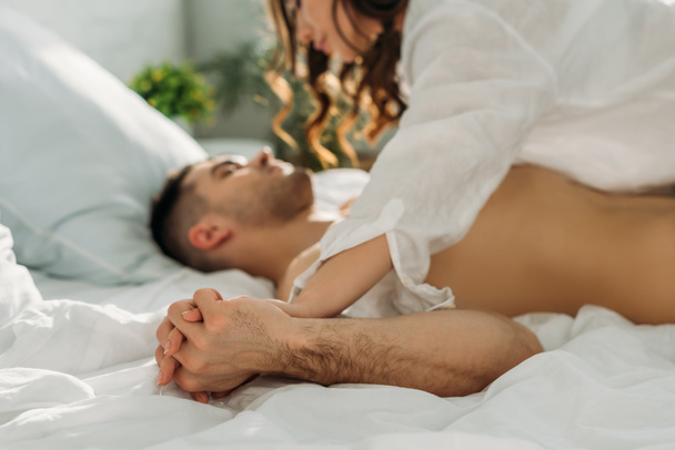 selective focus of seductive girl holding hands with sexy man while lying on him in bed - Fotografie, Obrázek
