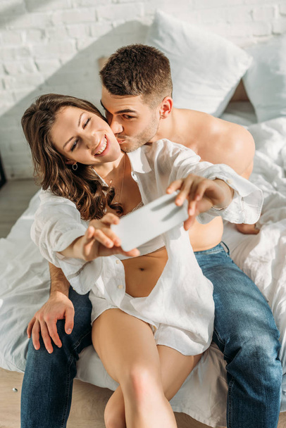 selective focus of happy girl taking selfie on smartphone while boyfriend kissing her - Foto, immagini