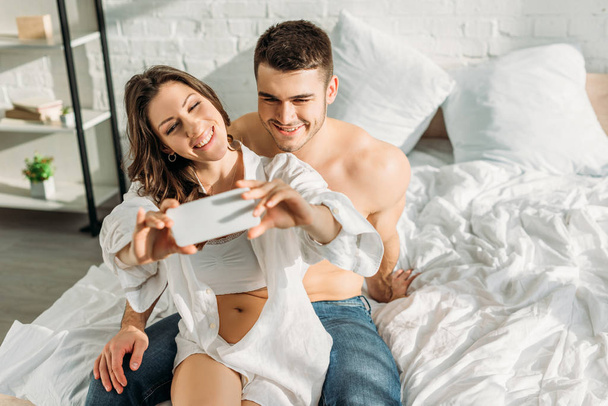 happy girl taking selfie on smartphone while sitting on bed with smiling boyfriend - Photo, Image