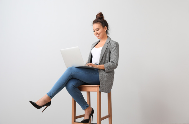 Young African-American businesswoman with laptop sitting on chair near grey wall - Foto, imagen