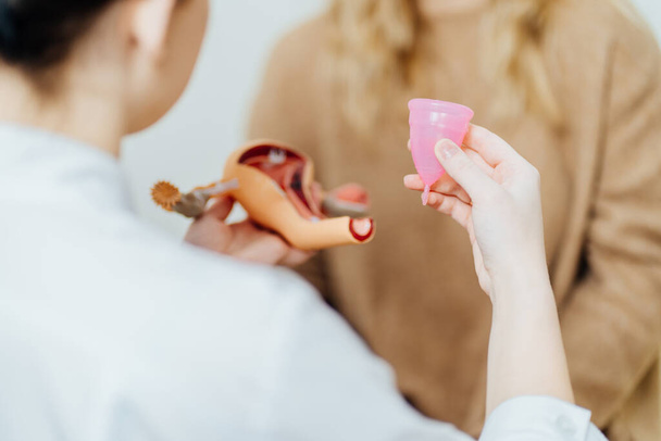 Doctor gynecologist holding anatomical model of uterus and menstrual cup, talking about intimate hygiene menstrual eco cup, how use, to female patient in an chair in modern medical office. - Photo, Image