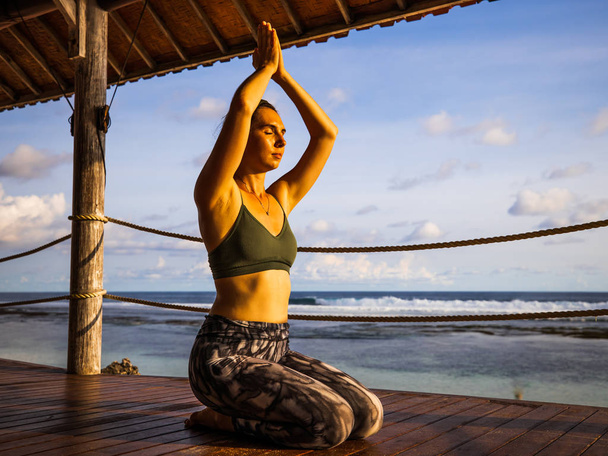 Caucasian woman practicing yoga, sitting on wooden floor. raising Arms with namaste mudra. Pranayama and meditation. Yoga retreat. Outdoor yoga practice. Melasti beach during sunset, Bali, Indonesia - Foto, Imagen