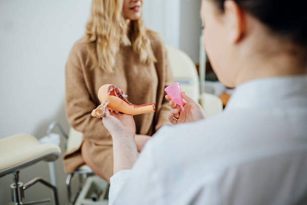 Γιατρός γυναικολόγος κρατώντας ανατομικό μοντέλο μήτρας και εμμηνορροϊκού κυπέλλου, μιλώντας για προσωπική υγιεινή έμμηνο οικολογικό κύπελλο, πώς χρήση, σε γυναίκα ασθενή σε μια καρέκλα στο σύγχρονο ιατρικό γραφείο. - Φωτογραφία, εικόνα