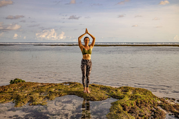 Yoga pose. Woman practicing yoga at the beach. Young woman standing and raising arms with namaste mudra. Healthy lifestyle. Yoga retreat in Bali, Melasti beach. - Photo, Image