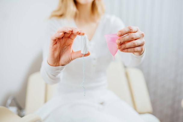 Young woman patient in gynecology chair holding menstrual cup and tampon and think what choosing. Question for doctor gynecologist. Consultation in the gynecological office. - Photo, Image