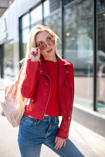 blonde in fashionable colored glasses looks away. Flirty girl Outdoor portrait of cute blonde lady wears elegant red jacket - Photo, Image