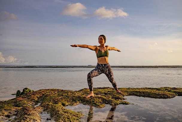 Молодая женщина практикует йогу, стоя в позе Вирабхадрасаны. Йога на открытом воздухе на пляже. Поза воина. Поработай. Здоровый образ жизни. Океан во время отлива. Йога, Бали
. - Фото, изображение