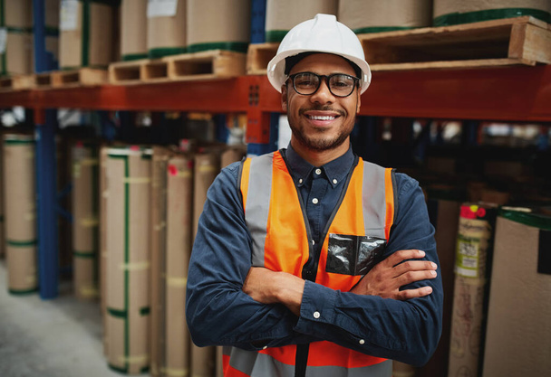 Portrait smiling worker wearing orange vest and white helmet with arms crossed in a large warehouse smiling - Fotoğraf, Görsel