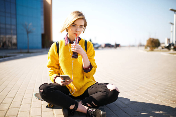 Young teenager girl sitting at the skatepark, holding mobile phone while listening to music with headphones.Pretty woman drinking from glass, with phone sitting on skateboard. Lifestyle, youth concept - 写真・画像