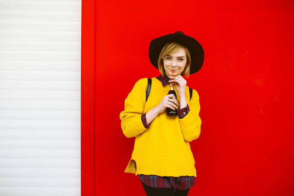 Young teenager girl model with drink posing over empty colorful red background. Hipster girl in a yellow sweater and hat drinking from glass and  doing emotion. Lifestyle, youth concept. - Photo, image