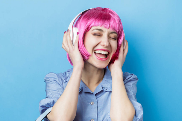 Chica feliz con peluca rosa escuchando música en línea con auriculares inalámbricos desde un teléfono inteligente aislado sobre fondo azul
 - Foto, Imagen