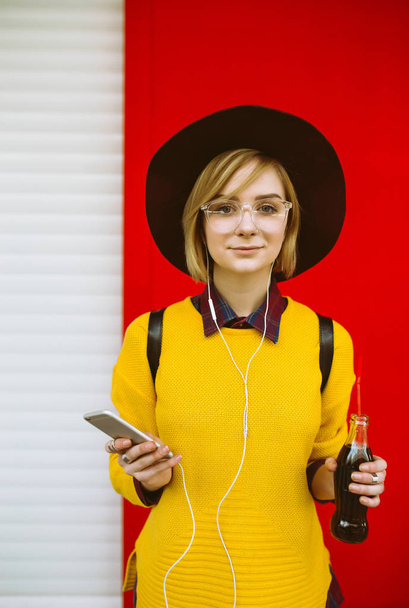Young teenager girl  with drink and phone posing on color background. Hipster girl in a yellow sweater and hat drinking from glass and texting on a mobile phone. Lifestyle, youth concept. - Foto, afbeelding
