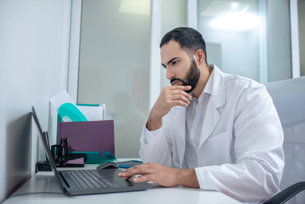 Male doctor in a white robe working at laptop looking serious - 写真・画像