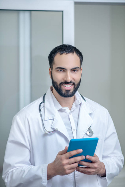 Male doctor in a white robe holding a tablet and smiling - Foto, Imagen