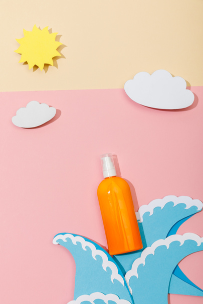 Top view of paper cut sea waves, clouds, sun and bottle of sunscreen on pink and beige  - Φωτογραφία, εικόνα