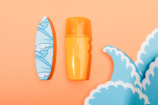 Top view of dispenser bottle of sunscreen with paper cut sea waves and surfboard on orange background - Foto, imagen
